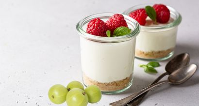 Zwei Gläser mit cremigem Dessert, verziert mit frischen Himbeeren und Minzblättern, daneben zwei Teelöffel und ein paar grüne Trauben auf einem weißen Untergrund