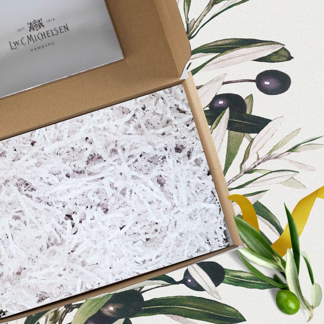 Geöffnete Geschenkbox aus braunem Karton mit weißem Papierfüllmaterial, dekoriert mit Olivenzweigen und gelbem Band auf floralem Hintergrund.