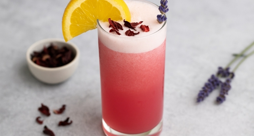 Ein rosa Hibiskus-Cocktail mit einer Orangenscheibe und Lavendelgarnitur, daneben eine Schale getrockneter Hibiskusblüten