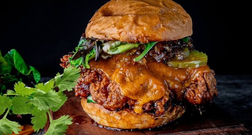 Ein Hähnchenburger mit geschmolzenem Cheddar, Gurkenscheiben und Salatblättern auf einem hölzernen Untergrund, dekoriert mit frischem Koriander.