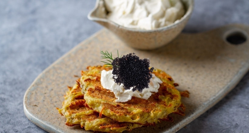 Zwei Kartoffelpuffer mit Creme Fraiche und schwarzem Kaviar, dekoriert mit Dill, serviert auf einem rustikalen Teller, im Hintergrund eine Schale mit Creme Fraiche