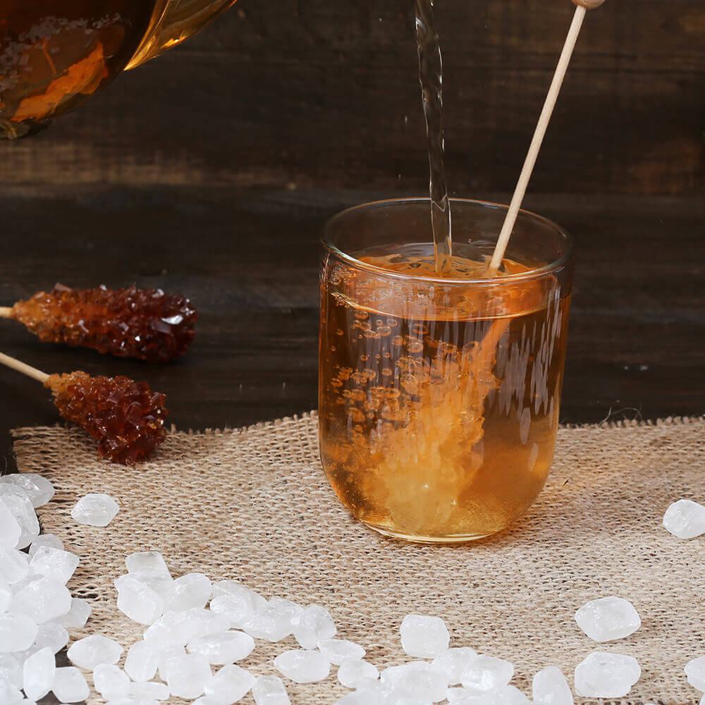 Bernsteinfarbenes Getränk wird in Glas mit Strohhalm eingegossen, umgeben von Kandiszucker und Zuckerlutscher auf Leinen.