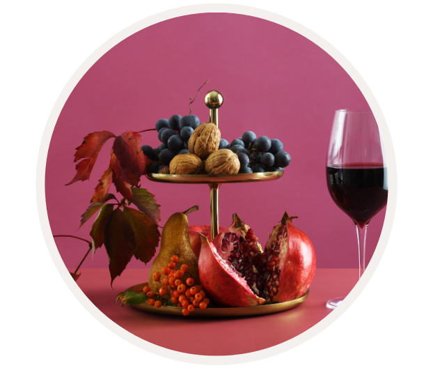 Ein Stillleben mit Granatapfel, Trauben, Nüssen, einer Birne und einem Glas Rotwein auf einem rosa Hintergrund.