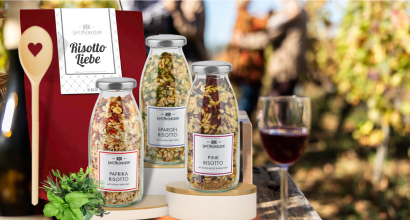 Risotto Geschenkset im Freien, mit Gläsern voller Paprika, Spargel und Pink Risotto, einem Holzlöffel und einem Glas Rotwein im Hintergrund