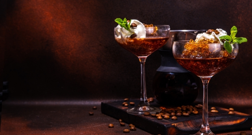 Zwei elegante Gläser mit Kaffeedessert, Sahnehaube und Minzblättern auf einem Holztisch mit Kaffeebohnen