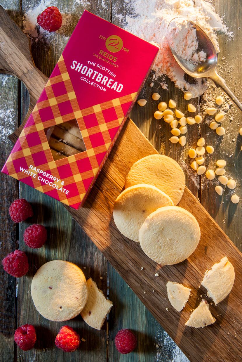 REIDS Shortbread Raspberry & White Chocolate Packung auf Holzuntergrund, daneben aufgeschnittene Shortbread-Kekse, Himbeeren und Perlen-Zucker.