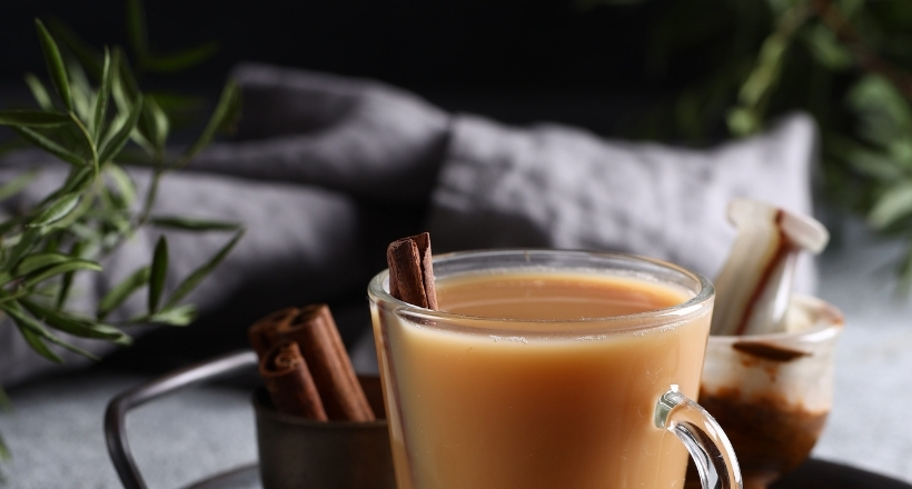 Ein Glas Chai-Latte mit Zimt und Gewürzen, auf einem Tisch serviert, umgeben von grünen Blättern