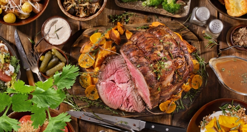 Ein saftiger, angebratener Fleischbraten, umgeben von Gewürzen und Beilagen auf einem Holztisch, garniert mit frischen Kräutern und Orangen.