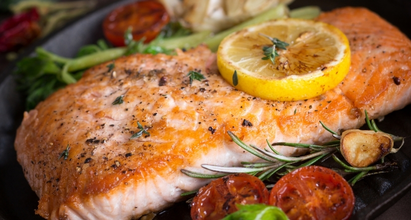 Nahaufnahme eines gegrillten Lachsfilets, garniert mit Zitronenscheibe, Rosmarin und gebackenen Tomaten.