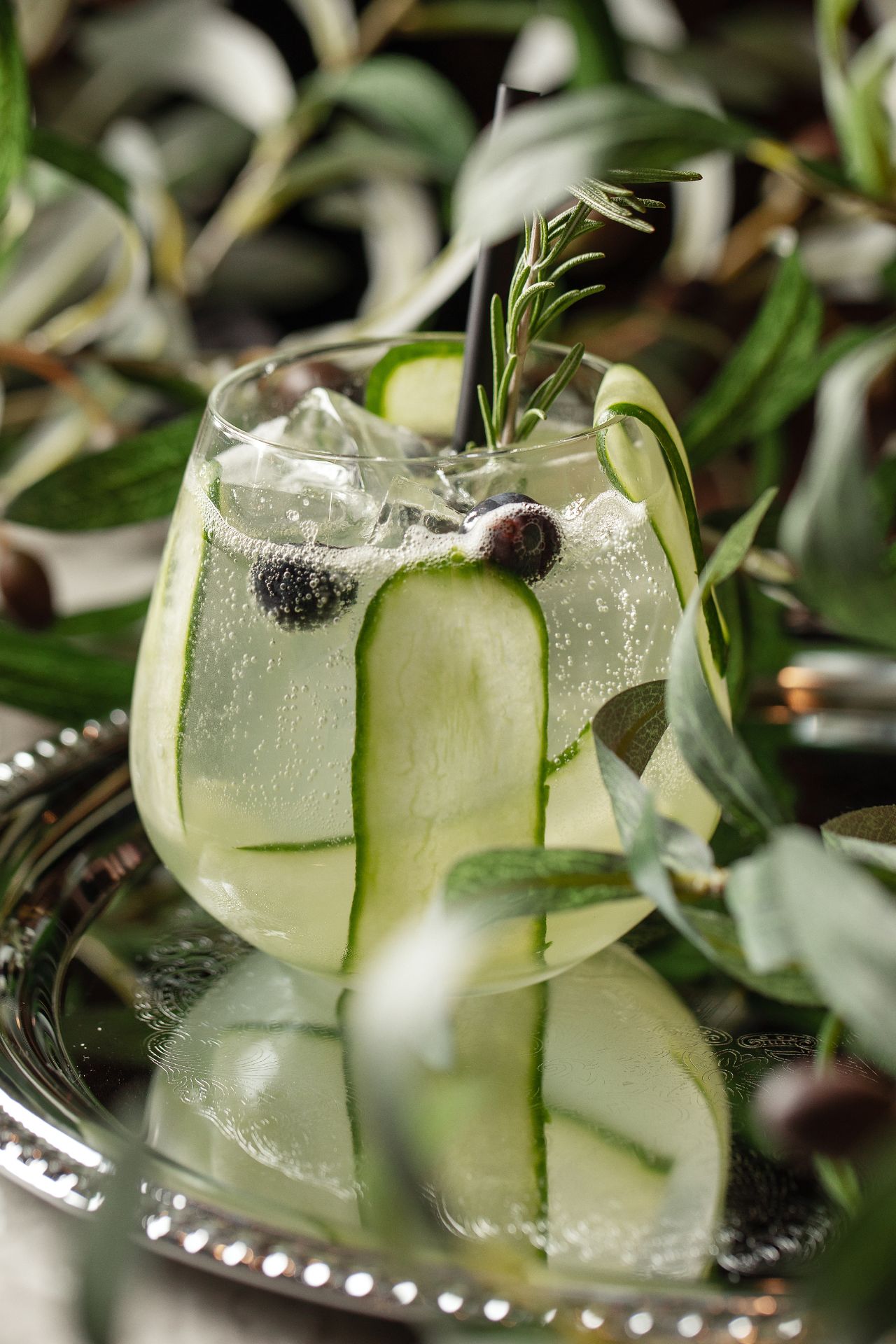 Ein Glas mit einem Cocktail, verziert mit Gurkenstreifen, Beeren und einem Zweig Rosmarin, serviert auf einem Tablett und umgeben von grünen Blättern.