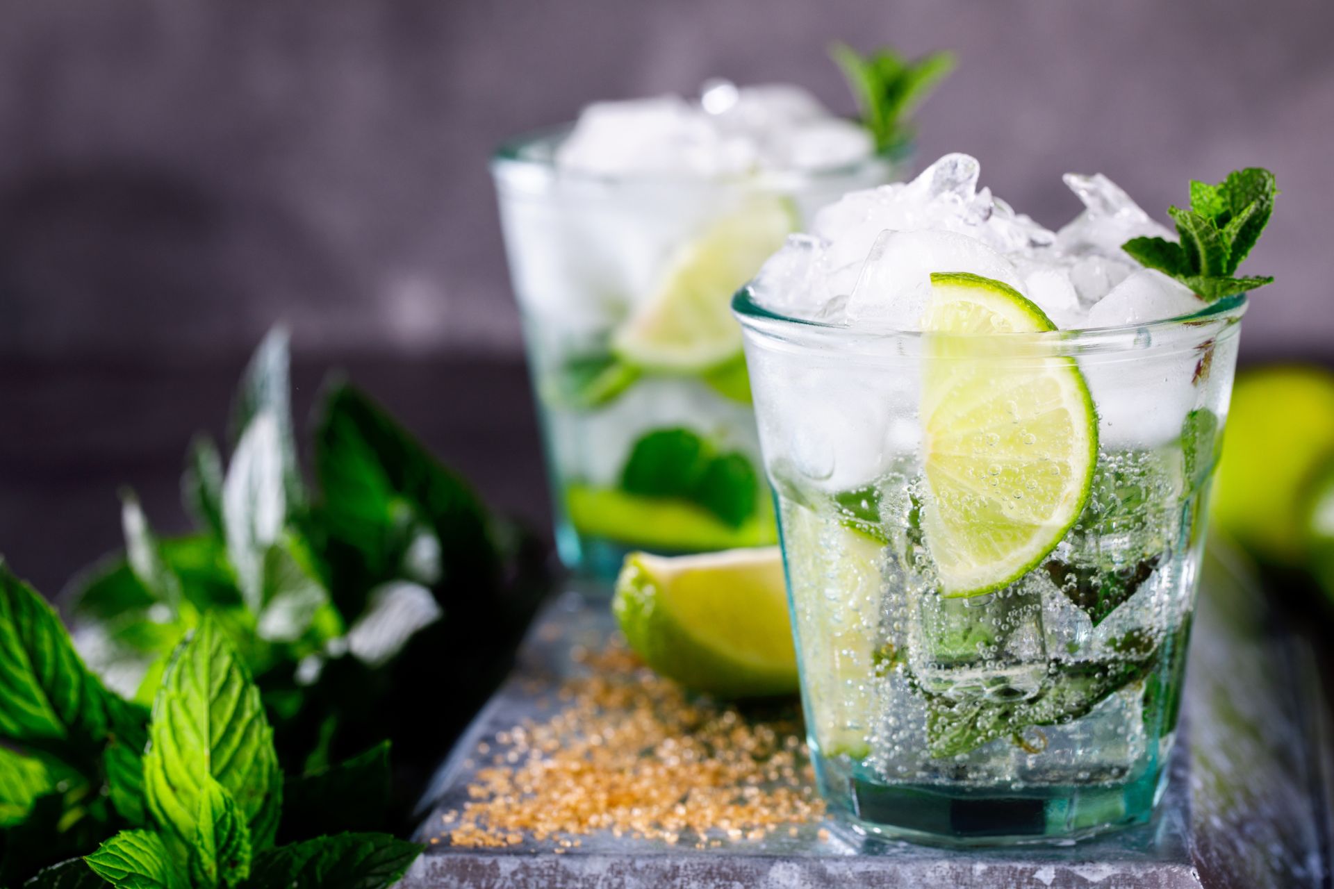 Nahaufnahme eines Mojito-Getränks mit Limettenscheiben, Minzblättern und Eiswürfeln in einem Glas, umgeben von frischen Minzblättern und braunem Zucker