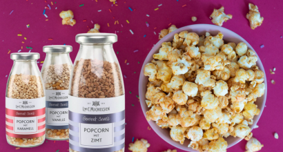 Drei Flaschen mit Popcorn-Gewürzen in den Geschmacksrichtungen Karamell, Vanille und Zimt neben einer Schüssel Popcorn auf pinkem Untergrund