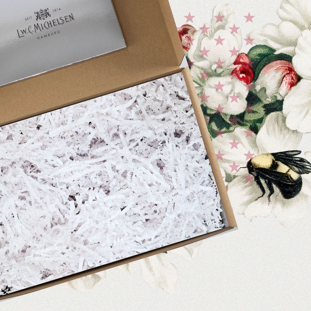 Geöffnete Geschenkbox aus braunem Karton, gefüllt mit weißem Papierfüllmaterial, daneben Nahaufnahme der floralen Banderole mit Bienenmotiv.