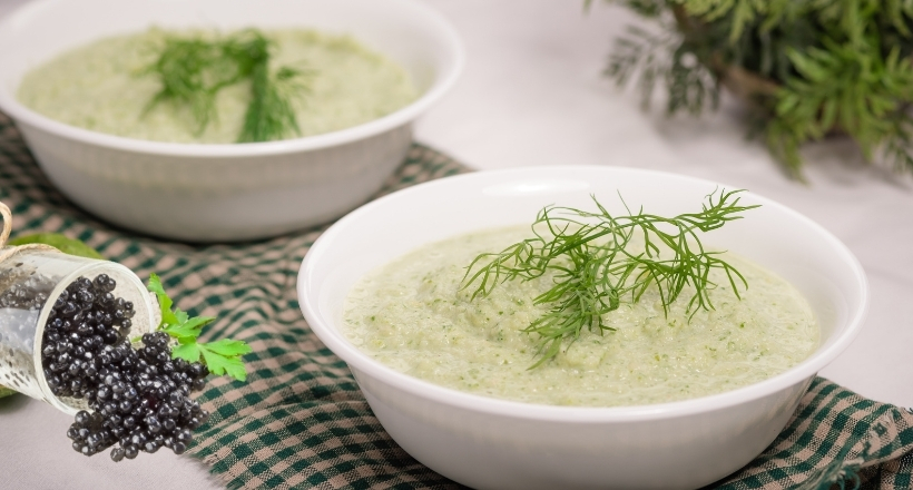 Zwei Schüsseln mit cremiger Gurkensuppe, garniert mit frischem Dill, auf einem karierten Tuch. Holunderbeeren liegen daneben.