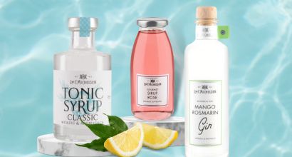 Flaschen mit Tonic-Sirup, Rosen-Sirup und Mango-Rosmarin Gin von Dr. Jaglas vor hellblauem Hintergrund mit Zitronenstücken