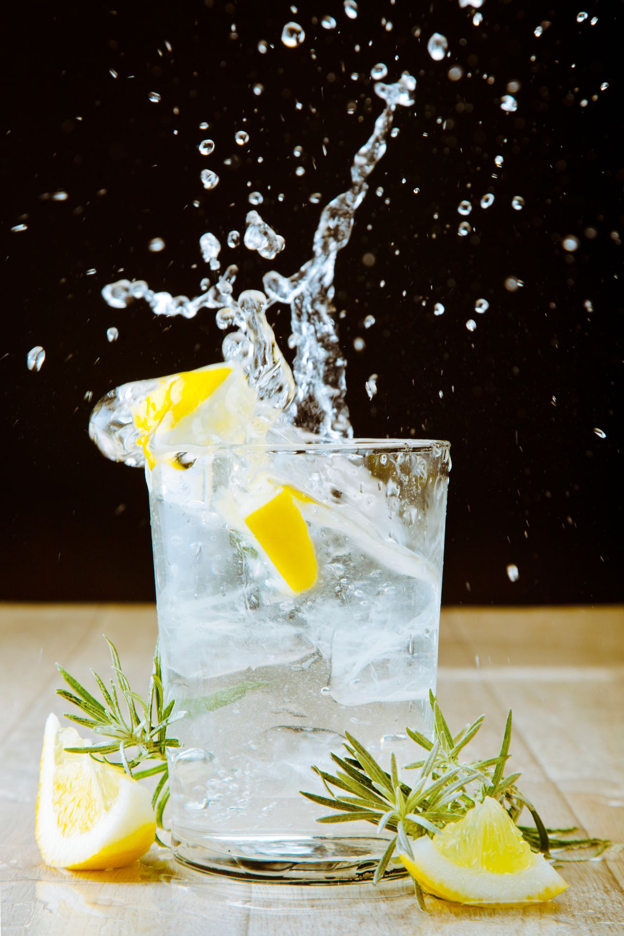 Ein Glas gefüllt mit eisgekühltem Wasser, Zitronenscheiben und Rosmarinzweigen, mit spritzenden Wassertropfen im Hintergrund