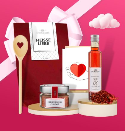 Ein Geschenkset bestehend aus einem Holzlöffel, mexikanischem Chilisalz, Chiliöl und Schokolade auf einem pinken Hintergrund, dekoriert mit Herzmotiv.