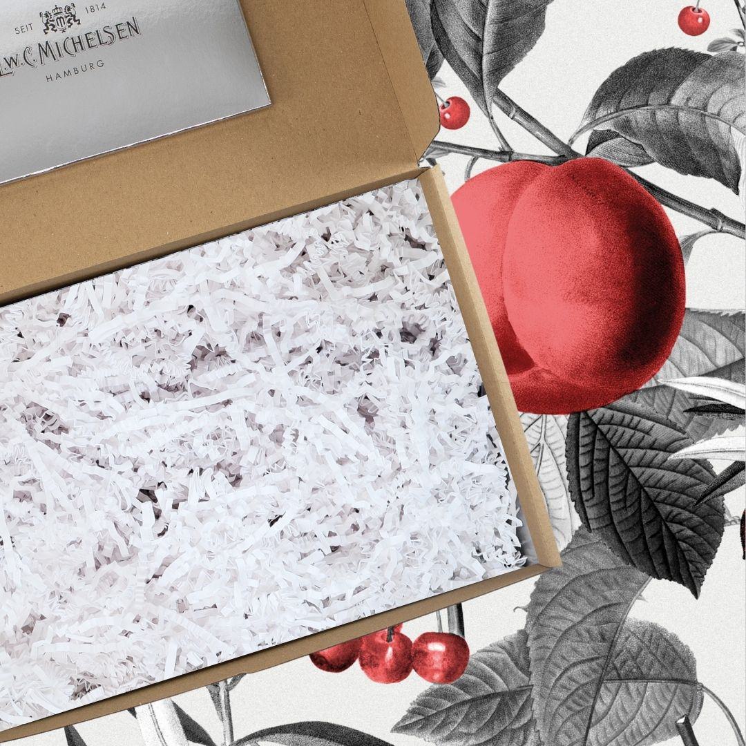 Geöffnete Geschenkbox aus braunem Karton mit weißem Papierfüllmaterial, daneben Detail des Musters mit roten Früchten und Blättern