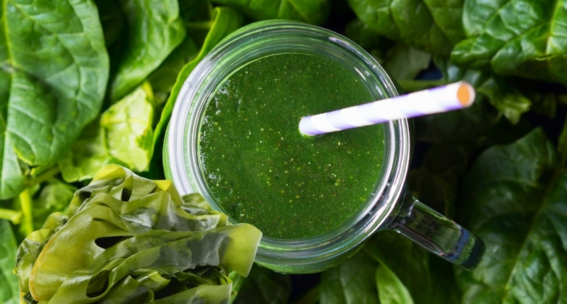 Glas mit grünem Smoothie und Strohhalm, umgeben von frischen Spinatblättern