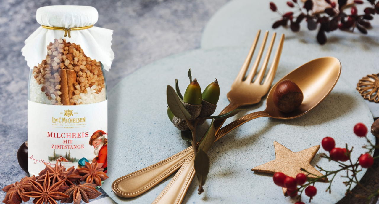 Flasche Milchreis mit Zimtstange von L.W.C. Michelsen neben goldenem Besteck auf einer festlichen Tischdekoration mit Nüssen und Gewürzen.