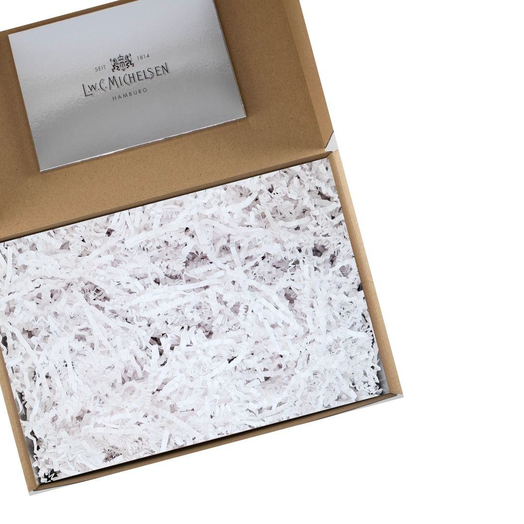 Geöffnete Geschenkbox aus braunem Karton mit weißem Papierfüllmaterial und silberner Karte im Deckel.