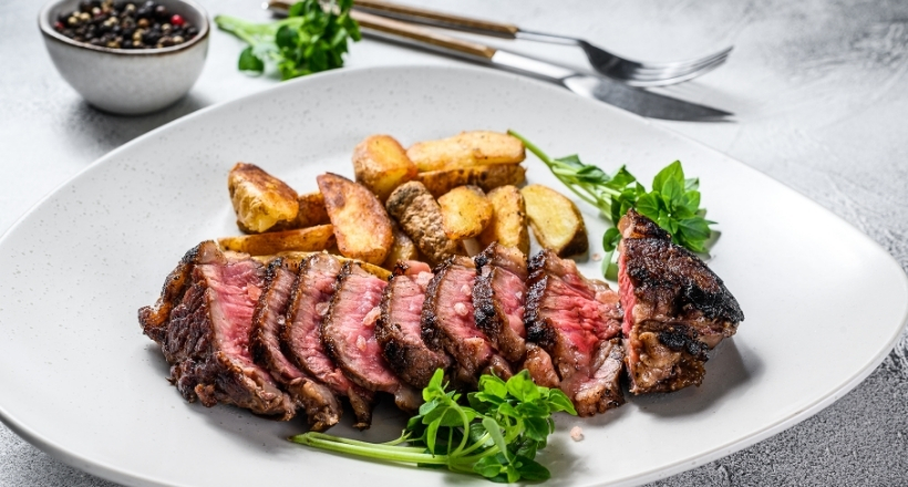 Ein saftiges, gegrilltes Steak serviert mit knusprigen Kartoffelecken und frischen Kräutern auf einem weißen Teller. Im Hintergrund befinden sich eine Schale mit Pfefferkörnern und Besteck.