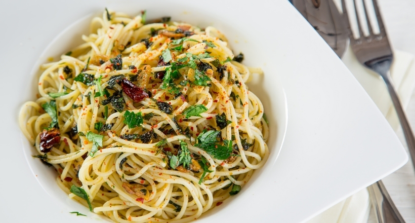 Ein Teller Spaghetti Aglio e Olio mit gehackten Kräutern und Chiliflocken, serviert in einer weißen Schale mit Gabel und Messer im Hintergrund.