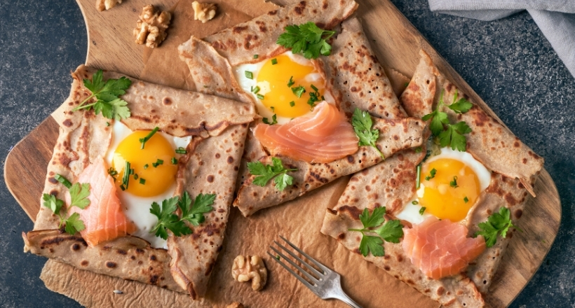 Drei herzhafte Galettes mit Lachs, Spiegelei, Petersilie und Schnittlauch auf einem Holzbrett serviert