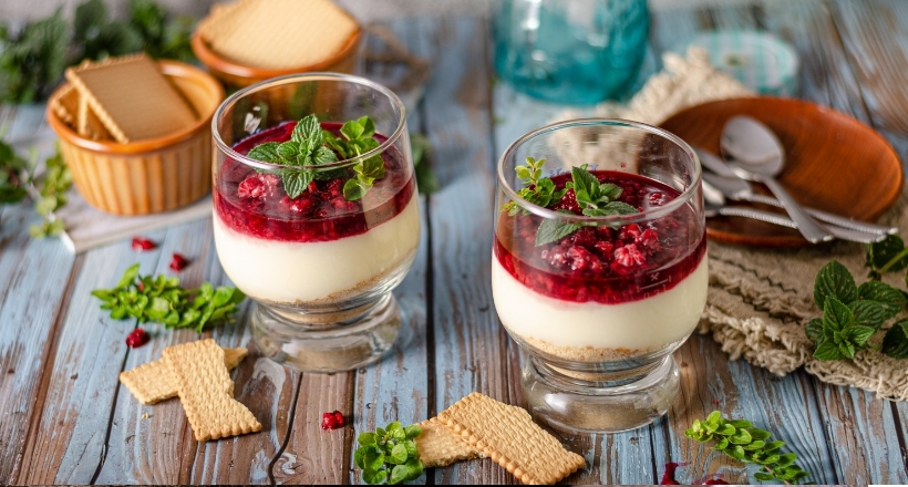Zwei Dessertgläser mit Schichten aus Creme, Himbeeren und Minze, dekoriert mit Keksen auf einem Holztisch