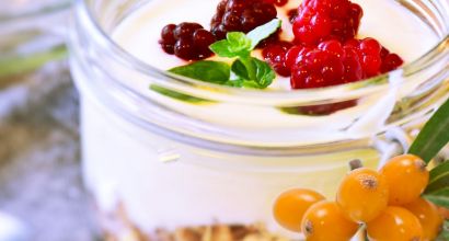 Ein Glas gefüllt mit Joghurt, dekoriert mit roten Beeren und grünen Kräutern, im Vordergrund ist Sanddorn zu sehen.