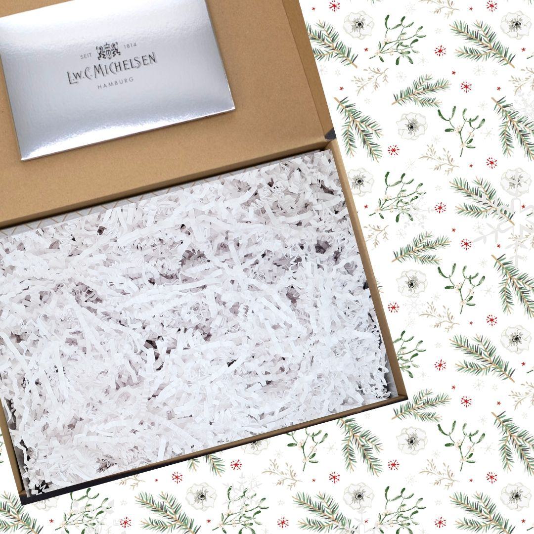 Geöffnete Geschenkbox aus braunem Karton mit weißem Papierfüllmaterial, daneben weihnachtliches Muster mit Tannenzweigen und floralen Elementen.