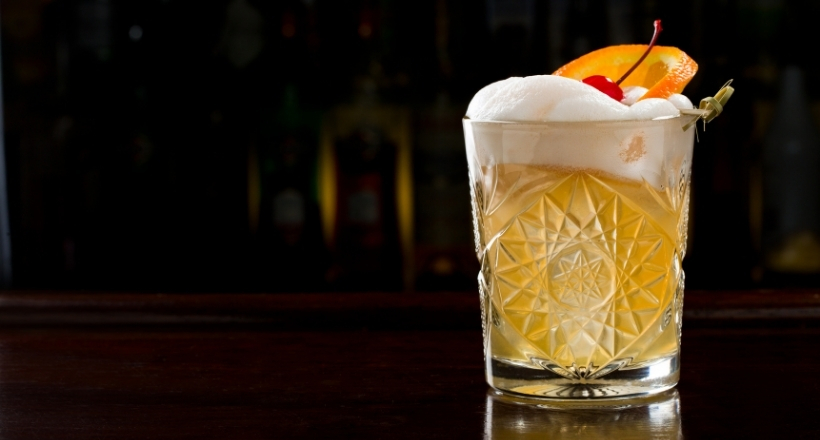 Ein kunstvoll gestalteter Whiskey Sour in einem Kristallglas, garniert mit einer Orangenscheibe und einer Kirsche, auf einem dunklen Barhintergrund.