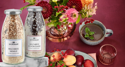 Zwei Flaschen Milchreis in den Sorten Vanille und Schokolade neben einer Blumenvase, einer Tasse, einer Obstschale und einem Glas auf einem Tisch