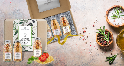 Geschenkbox mit drei Flaschen Dinner-Mischungen, dekoriert mit frischen Kräutern und Gewürzen auf hellem Hintergrund