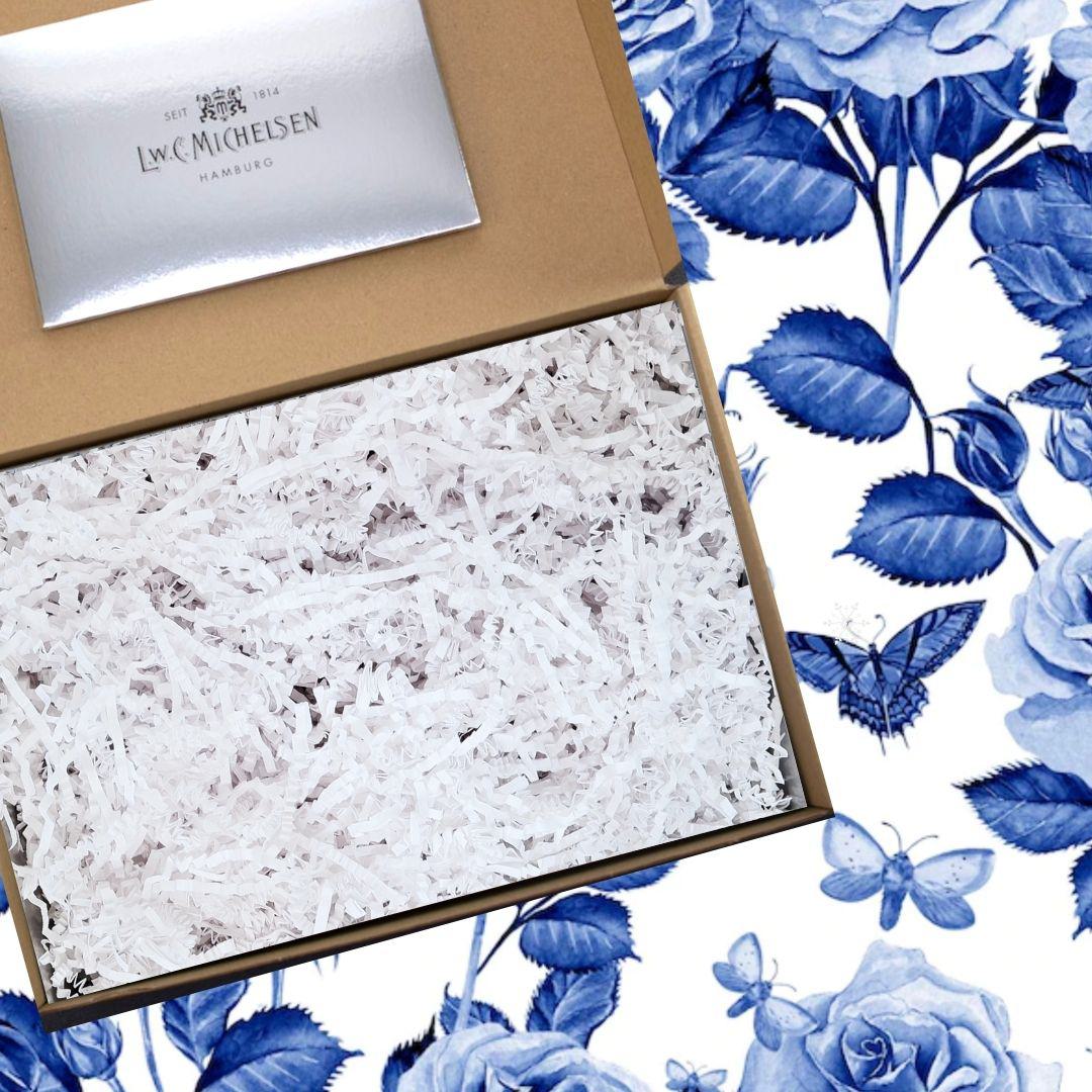 Geöffnete Geschenkbox aus braunem Karton mit weißem Papierfüllmaterial, daneben Detail des blauen floralen Musters mit Rosen und Schmetterlingen