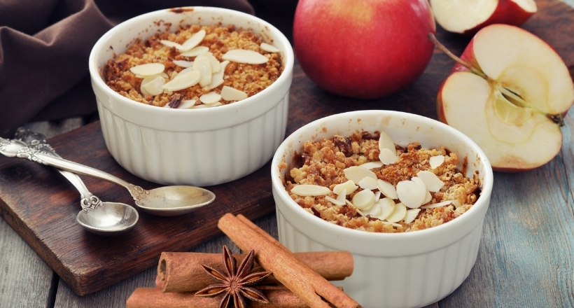 Zwei Schalen mit Apfel-Crumble, garniert mit Mandeln, im Hintergrund frische Äpfel und Zimtstangen