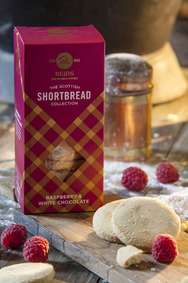 Karton REIDS Scottish Shortbread Collection Raspberry & White Chocolate auf Holzbrett mit ausgepackten Keksen und frischen Himbeeren, im Hintergrund Glas mit Getränk.