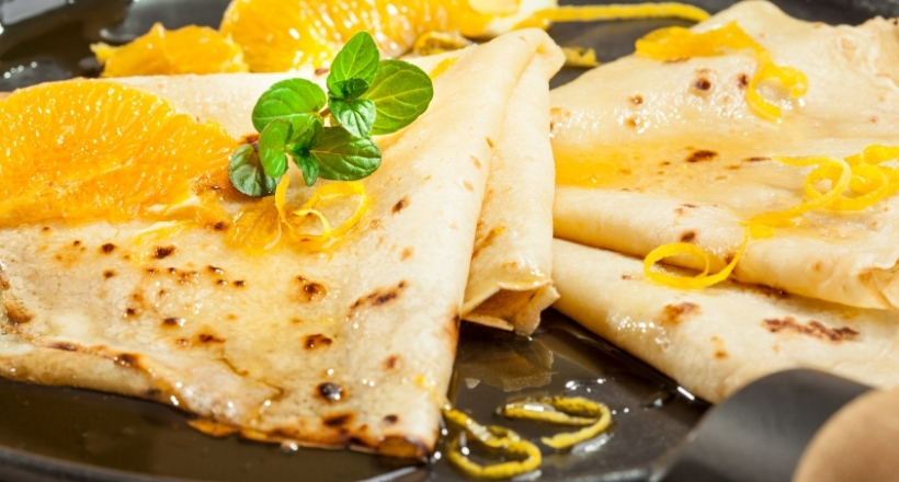 Nahaufnahme von goldenen Crêpes, garniert mit frischen Orangenscheiben, Orangenabrieb und einem Zweig Minze auf einem Teller.