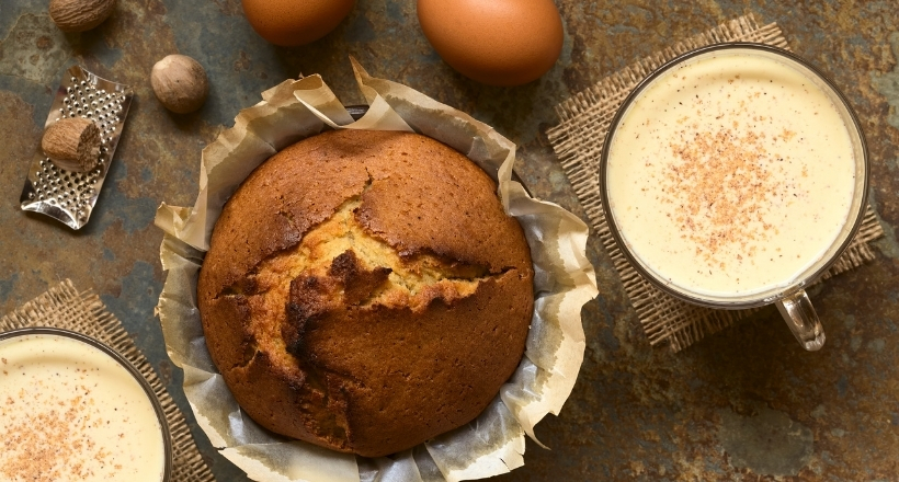 Ein frisch gebackener Kuchen auf Backpapier, umgeben von Eiern, Muskatnüssen und einer Tasse würzigen Getränks auf einem rustikalen Tisch